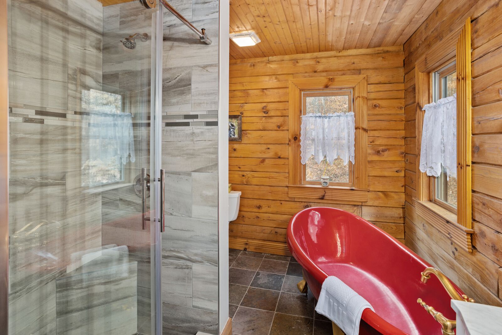 Une salle de bain rustique, avec des murs en bois, une douche en verre, une baignoire rouge et une fenêtre agrémentée d’un rideau.