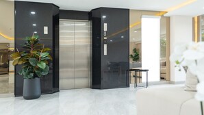 A modern lobby with a marble floor, a large potted plant, a reception desk, and a glass elevator. - Oriole Residence (Bang Phli)
