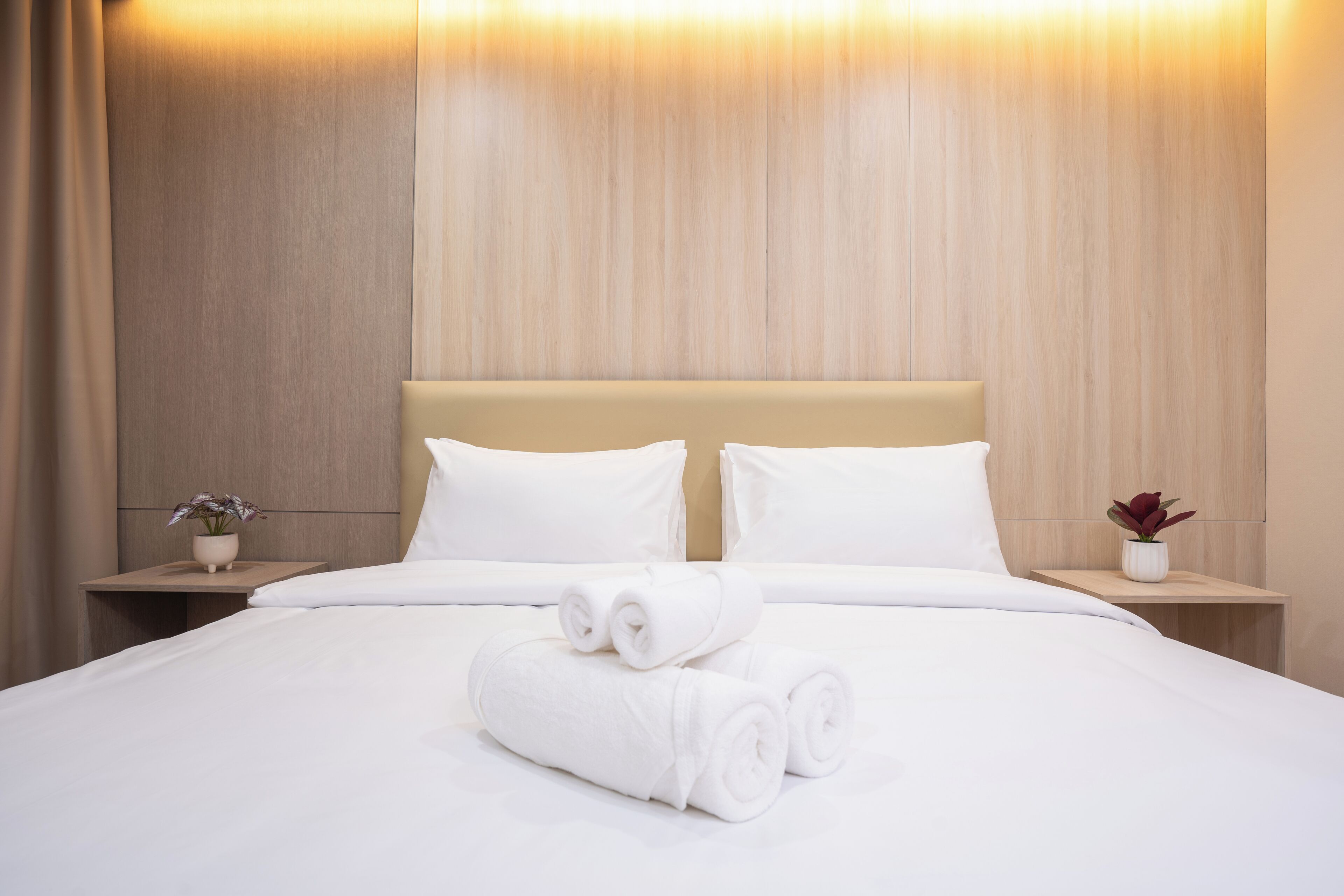 A neatly made bed with white linens and rolled towels, two bedside tables with potted plants, and a wooden headboard.