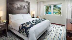 Habitación de hotel con una cama grande, una mesita de noche con lámpara, una silla y vista a una playa a través de una ventana.