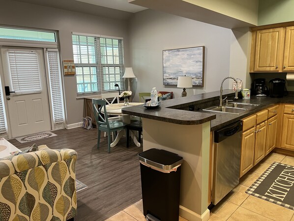 A modern kitchen with a central island, wooden cabinets, and a dining area with a table and chairs. - 3BR Beach Condo Walk to Shops & Restaurants (Pensacola)
