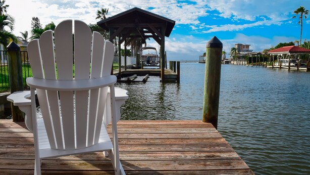 Una banchina in legno con una poltrona Adirondack bianca, un'area ombreggiata e vista sull'acqua e edifici in lontananza.