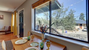 Uma área de refeições com mesa posta para quatro pessoas, uma janela com vista para árvores e um edifício, e uma cadeira com almofada.