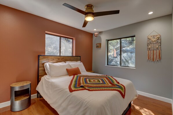 A bedroom with a wooden headboard, a bed with white linens, a colorful blanket, a ceiling fan, and a window with a view of trees.