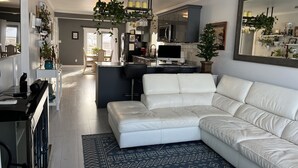 Ein modernes Wohnzimmer mit einem weißen Ledersofa, einem Essbereich mit Tisch und Stühlen, einer Küche mit Kühlschrank und Fernseher sowie einem Deckenventilator.