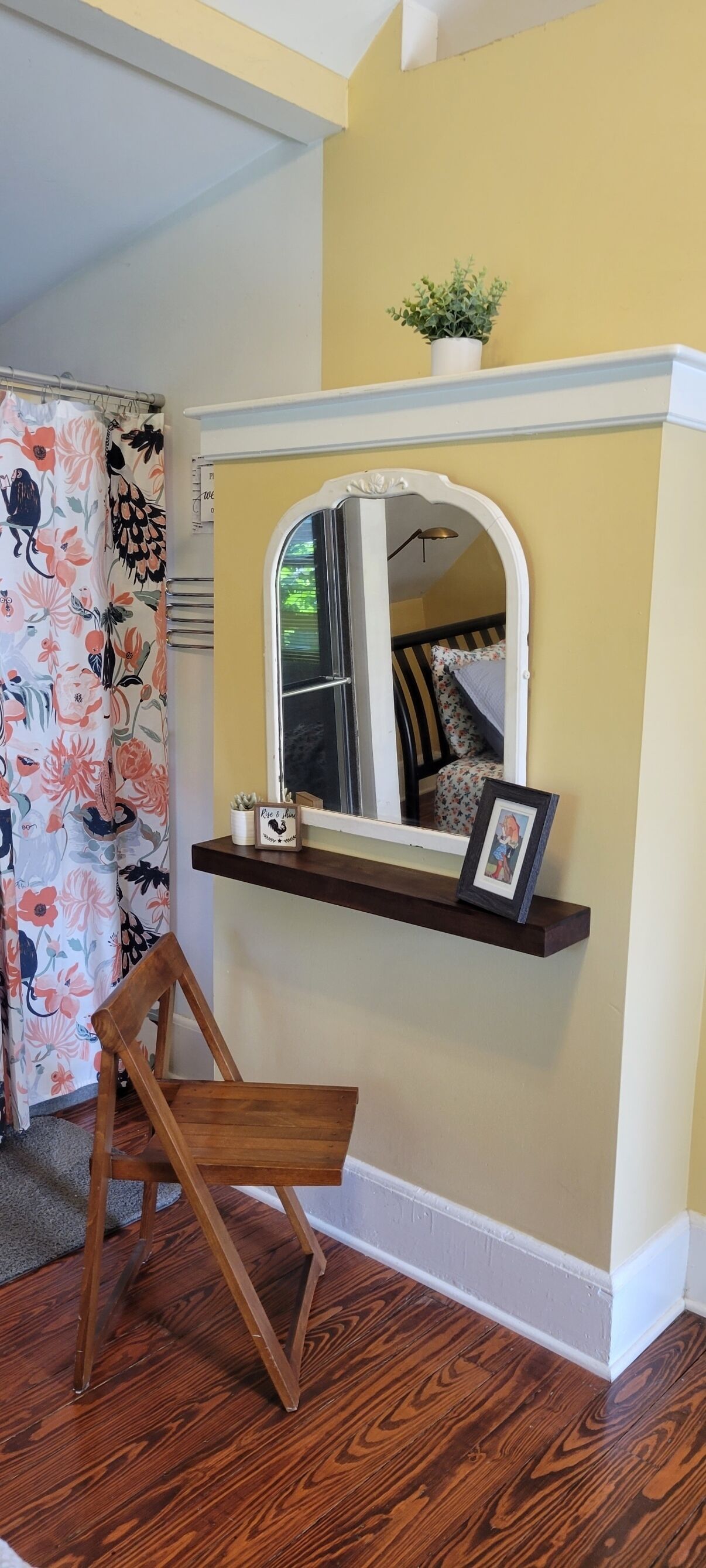 A room with a wooden chair, a small shelf with a framed picture, a potted plant, and a mirror above the shelf.