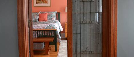 Une pièce dotée d’une porte à cadre en bois qui donne sur une chambre. La pièce a un mur gris, un plancher en bois et un lit avec une literie à motifs floraux.