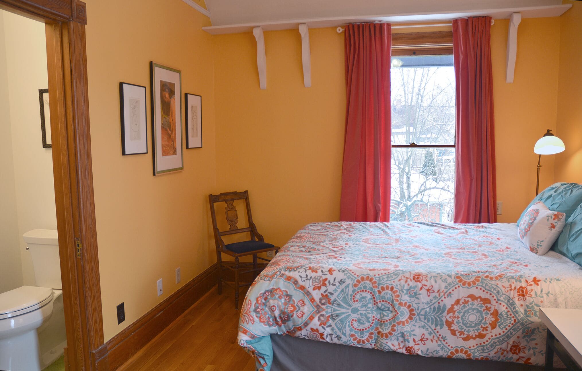 A bedroom with a bed, a chair, a window with curtains, and framed pictures on the wall.