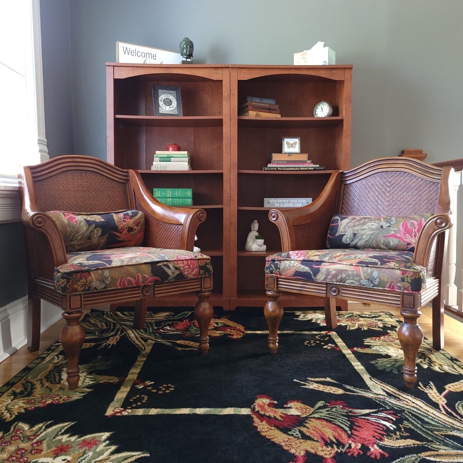 Quarto com duas poltronas estofadas com tecido floral, uma estante de madeira com diversos objetos e um tapete escuro com padrão de galo.