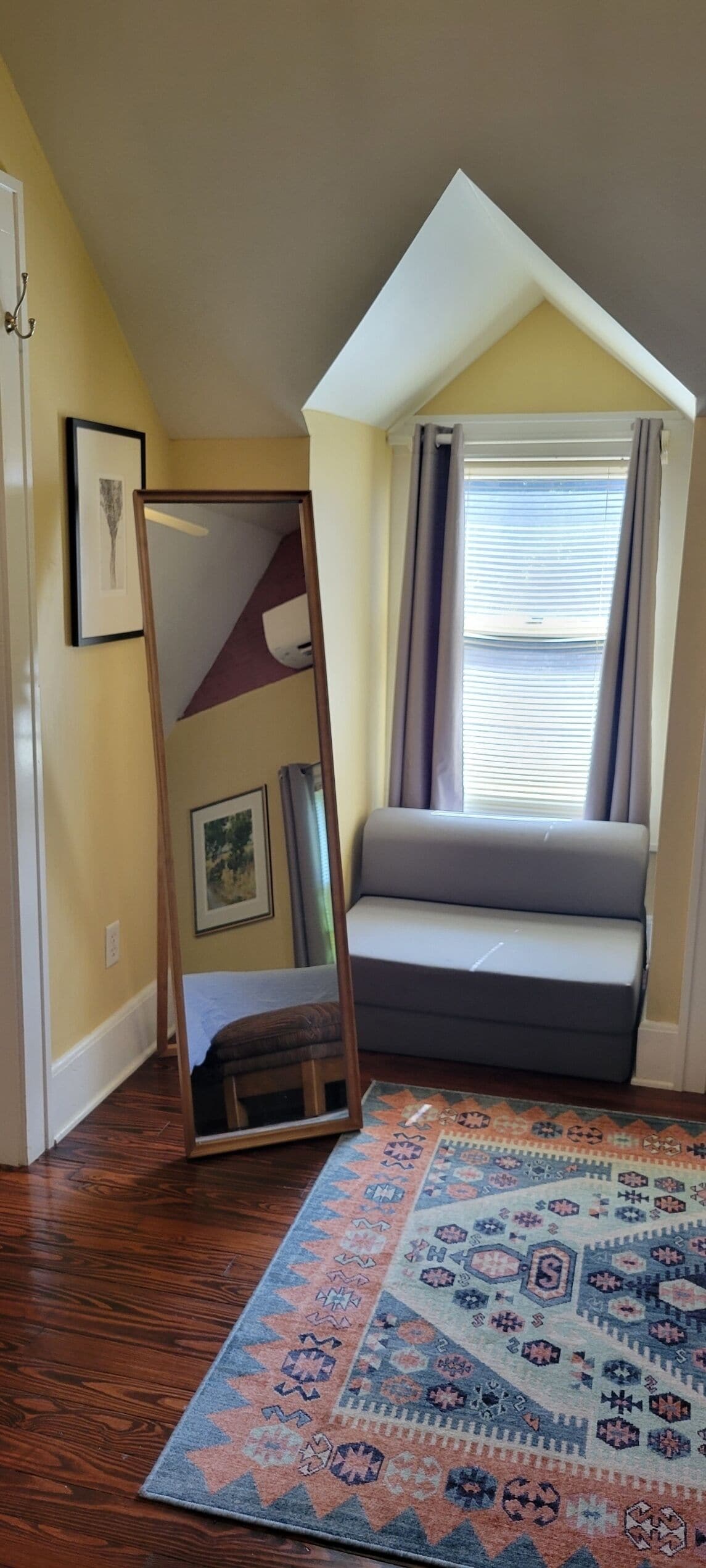 Una habitación con techo inclinado, una ventana con cortinas, un sofá, un espejo y una alfombra estampada.