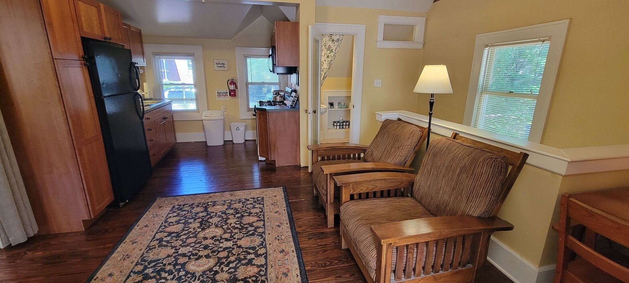 A spacious living area with a wooden sofa set, a patterned rug, and a kitchen in the background.