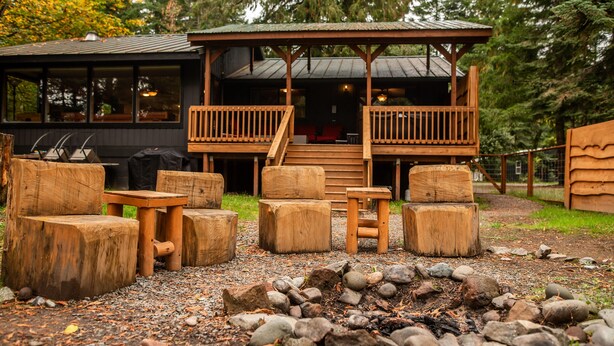 Una terraza de madera con mobiliario exterior y una chimenea, rodeada por un edificio de estilo rústico tipo cabaña.