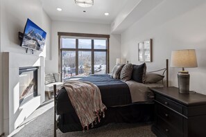 A modern bedroom with a large window, a fireplace, a bed with a blanket, and a bedside table with a lamp.