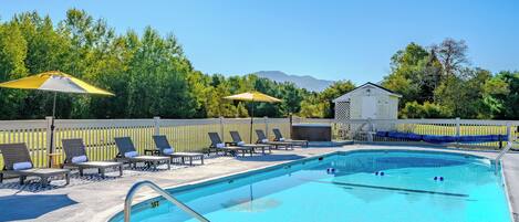 A swimming pool with sun loungers and umbrellas, surrounded by a wooden fence and trees.