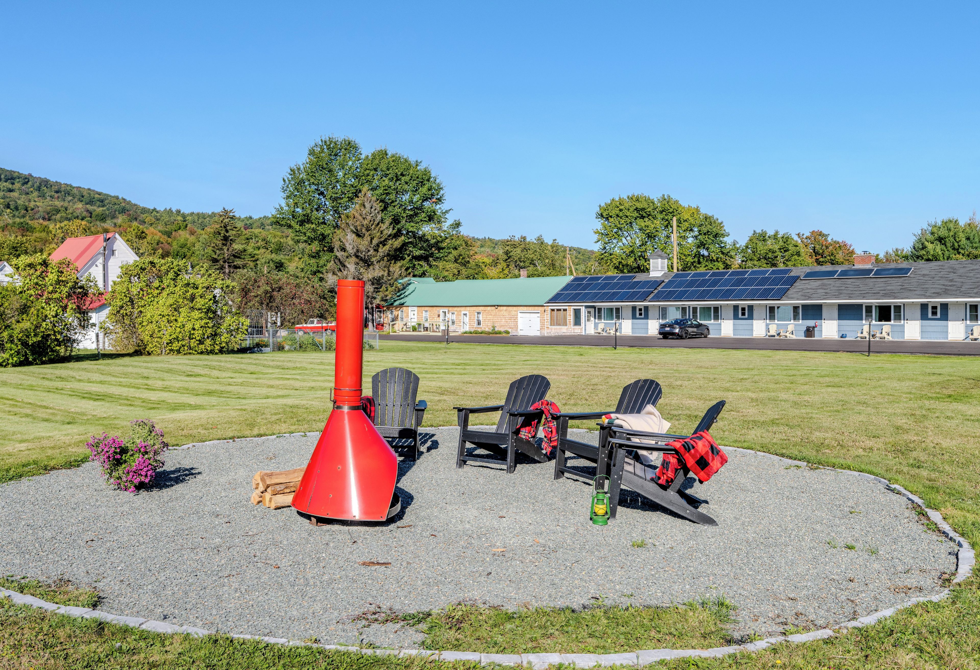 A circular outdoor seating area with Adirondack chairs, a red fire pit, and a gravel path.