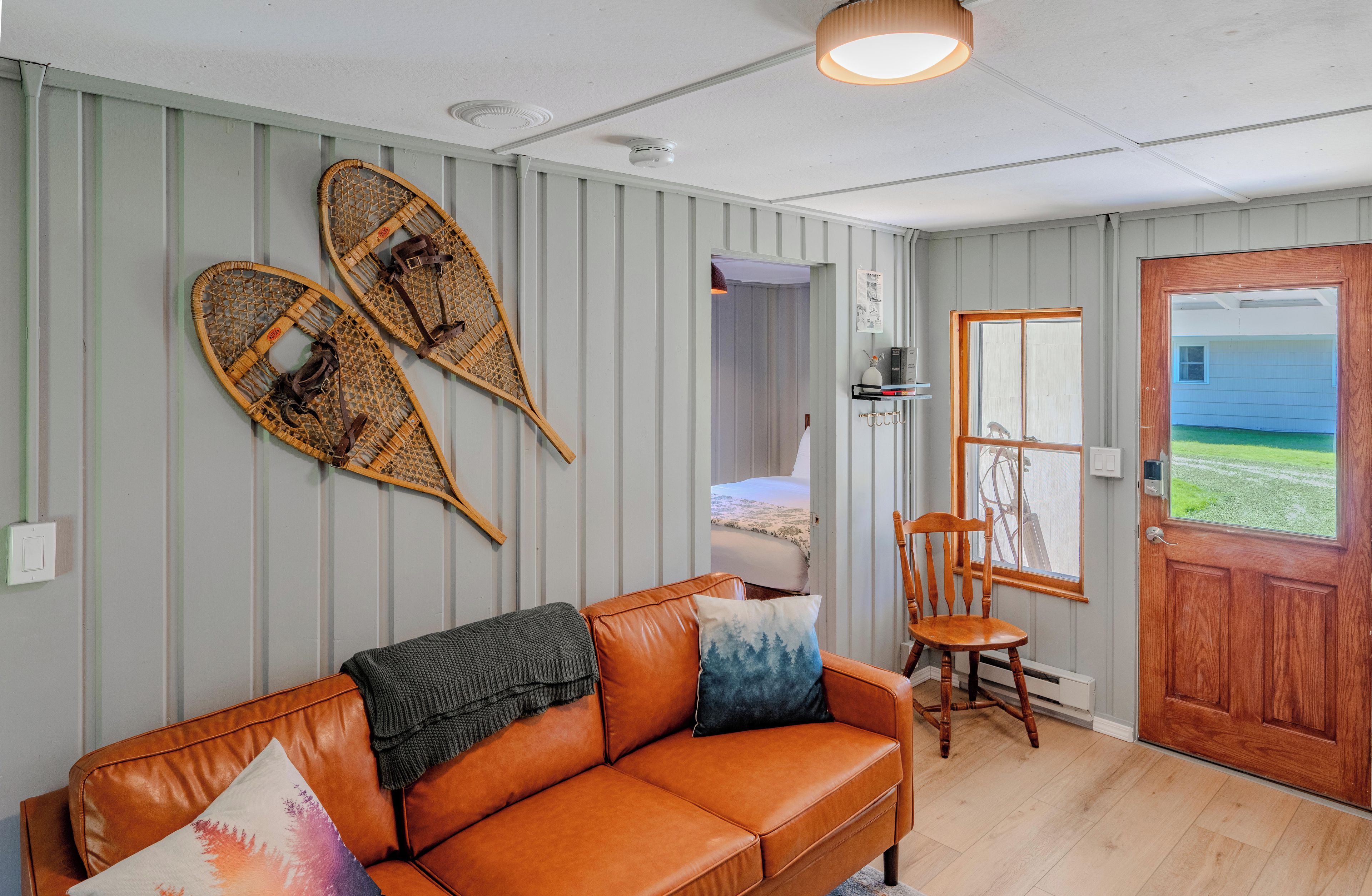 Una habitación con un sofá de cuero marrón, una silla de madera, una puerta con vistas y dos objetos fijados a la pared.