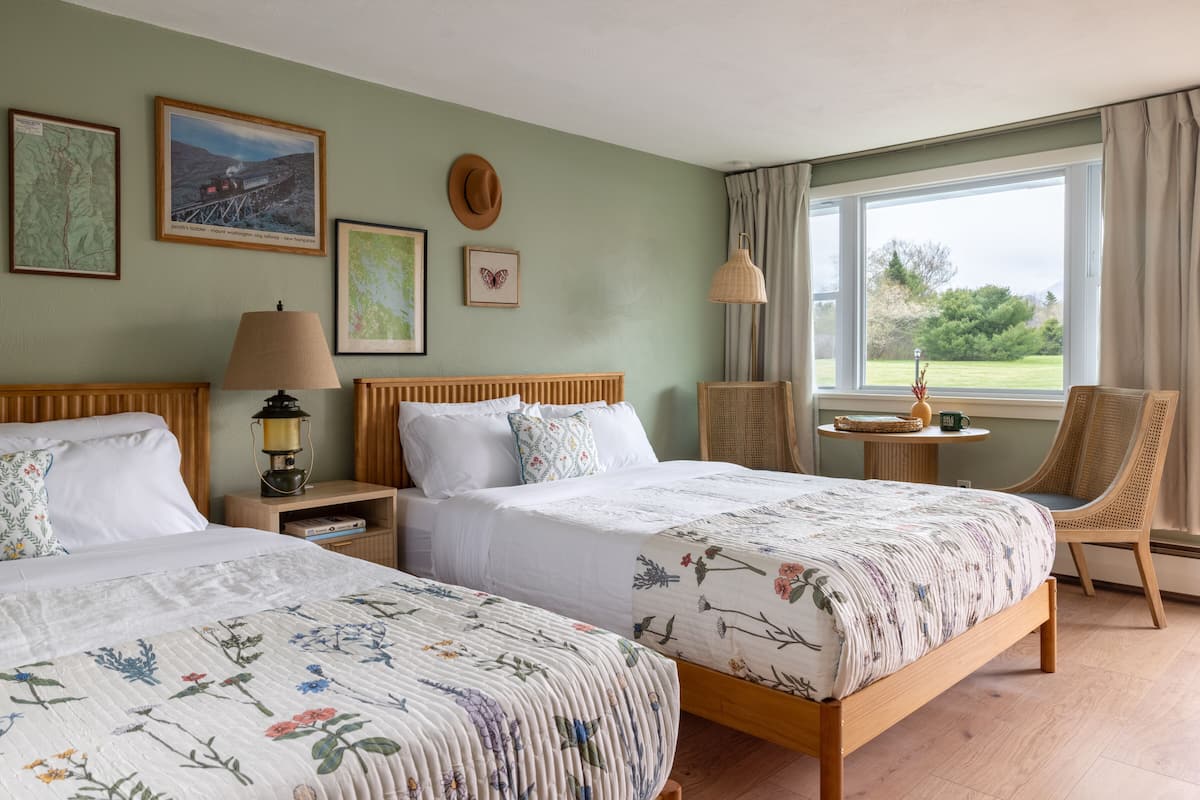 A hotel room with two beds, a wooden headboard, a nightstand with a lamp, a small table with a vase, and a window with a view of greenery.
