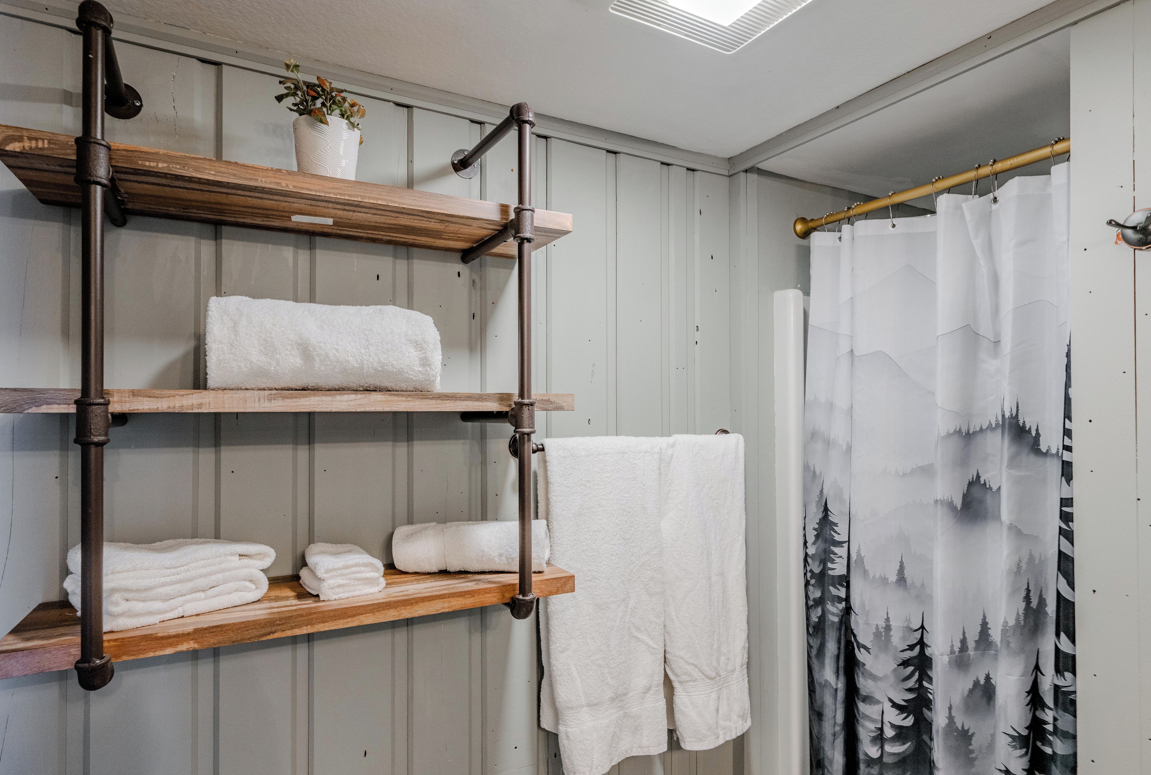 A bathroom with wooden shelves holding towels, a shower curtain with a forest design, and a potted plant on the top shelf.
