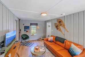 A modern living room with a leather sofa, a television, a wooden coffee table, and a large window with a view of mountains. - Gale River House - Boutique Hotel (Franconia)