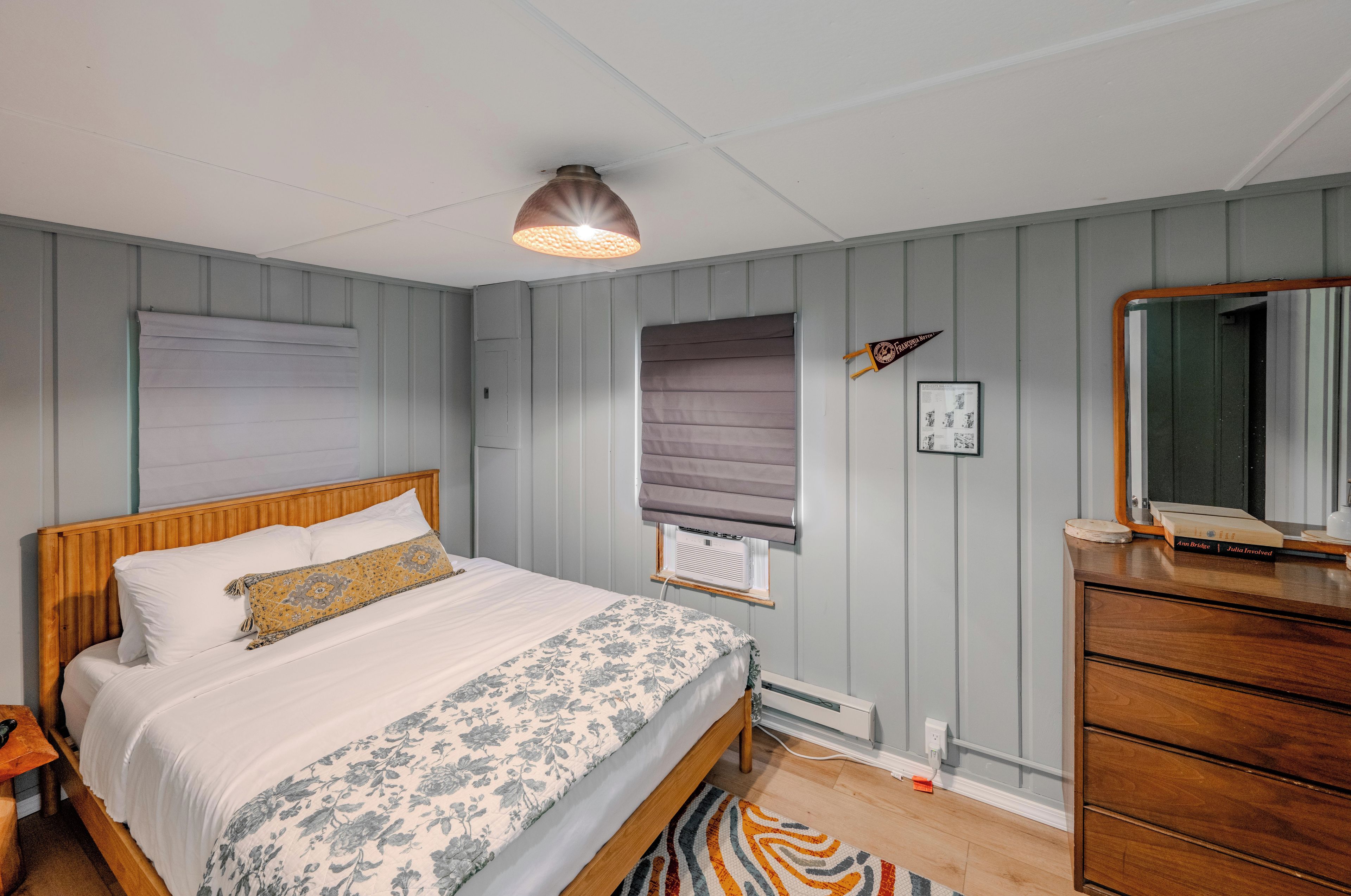 A bedroom with a wooden bed, a bedside table, a dresser, a radiator, and a window with blinds.