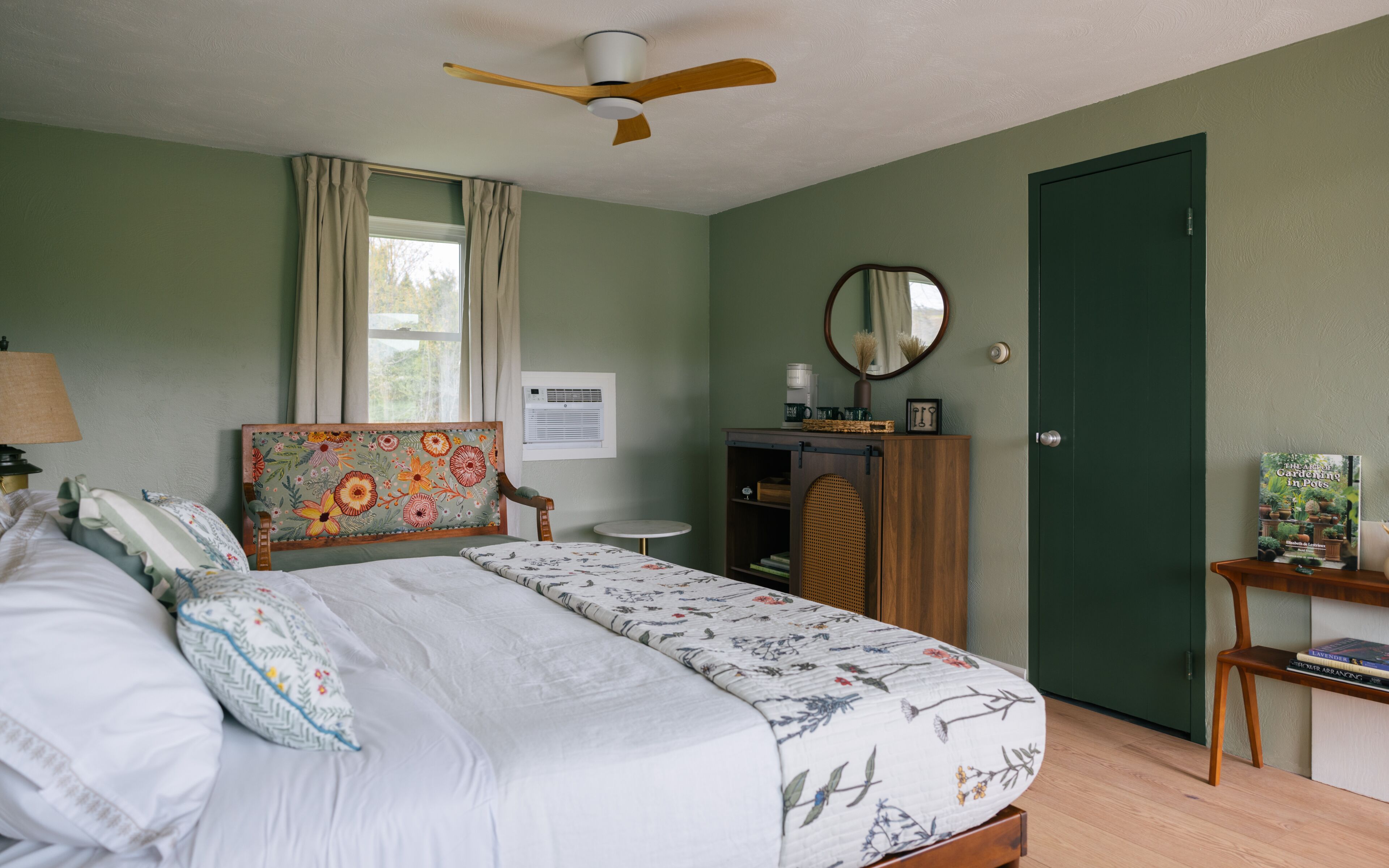 A bedroom with a bed, a ceiling fan, a window with curtains, a bedside table, a dresser, and a door.