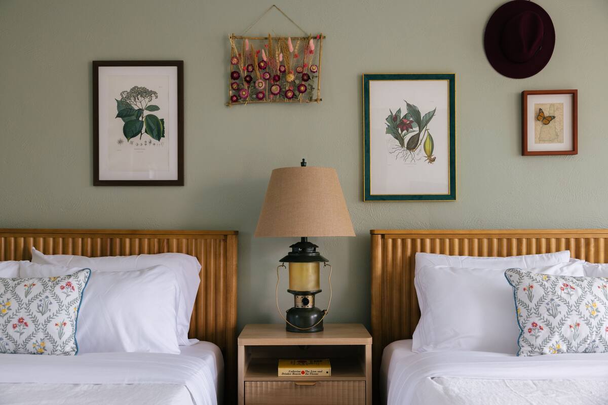 A hotel room with two beds, a nightstand with a lamp, and framed botanical illustrations on the wall.