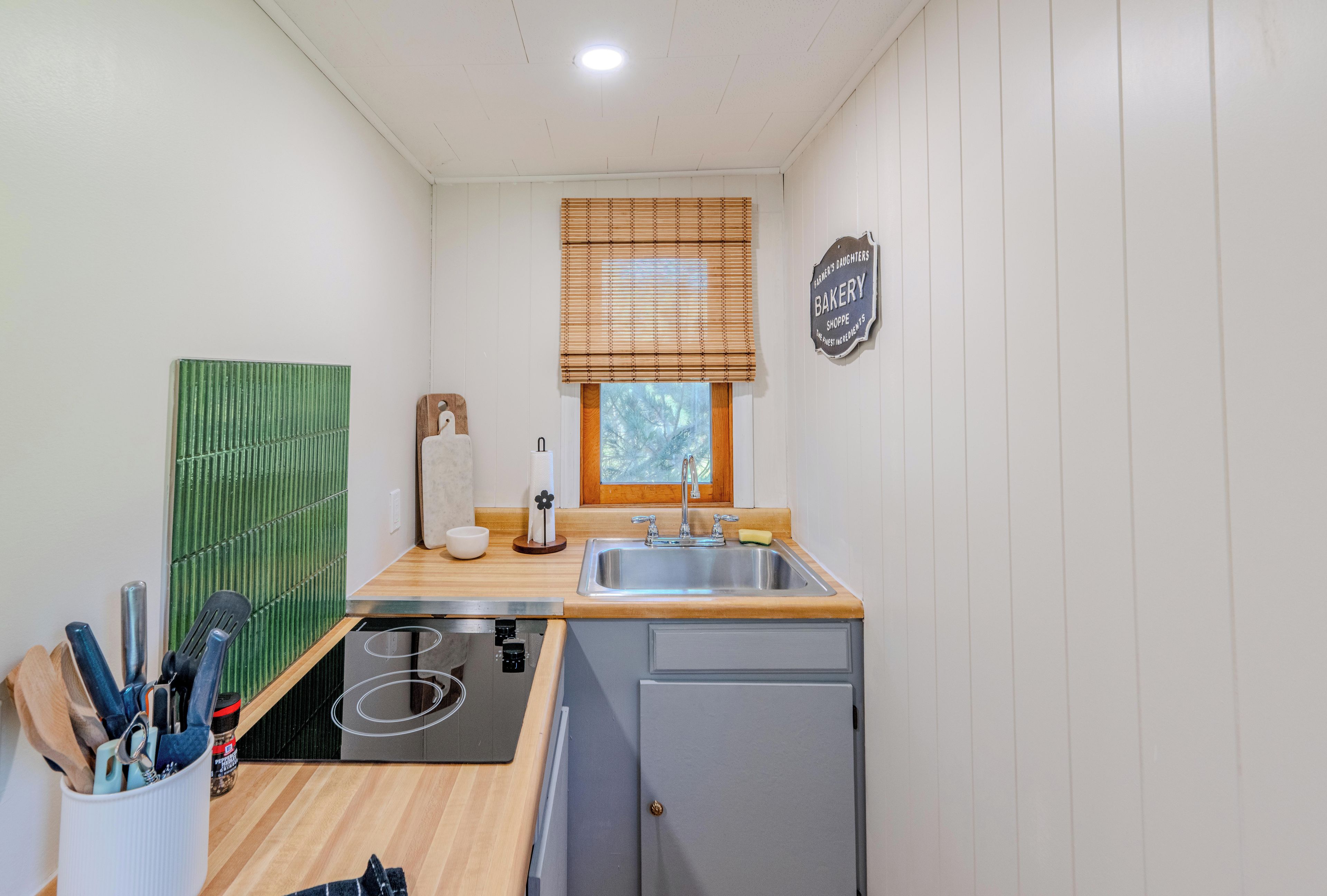 A compact kitchen with a sink, a window with blinds, a small refrigerator, and a green backsplash.