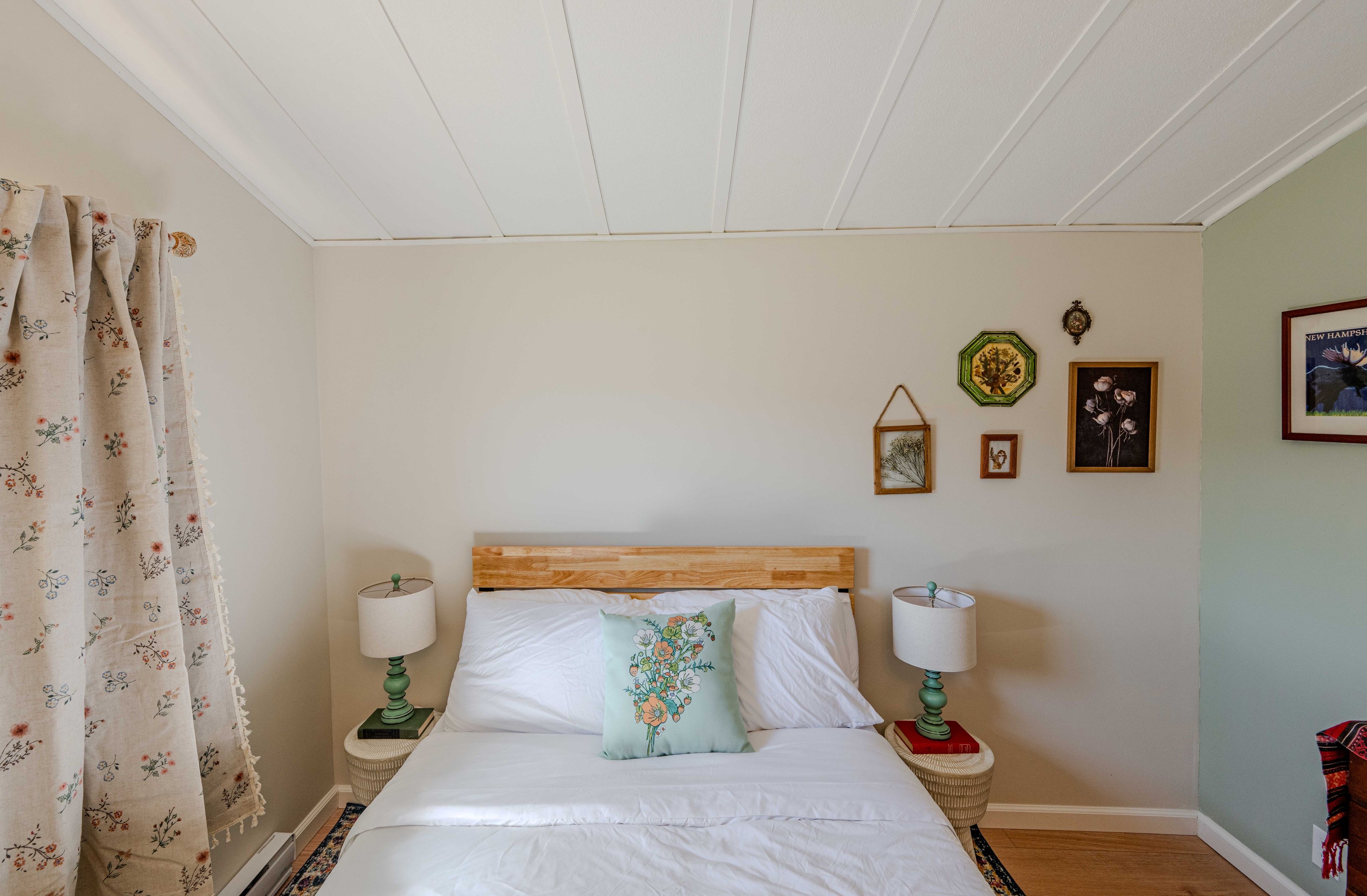 Une chambre à coucher avec une tête de lit en bois, une literie blanche, deux lampes de chevet, un coussin à motifs floraux et un lit sur lequel se trouve un livre rouge.