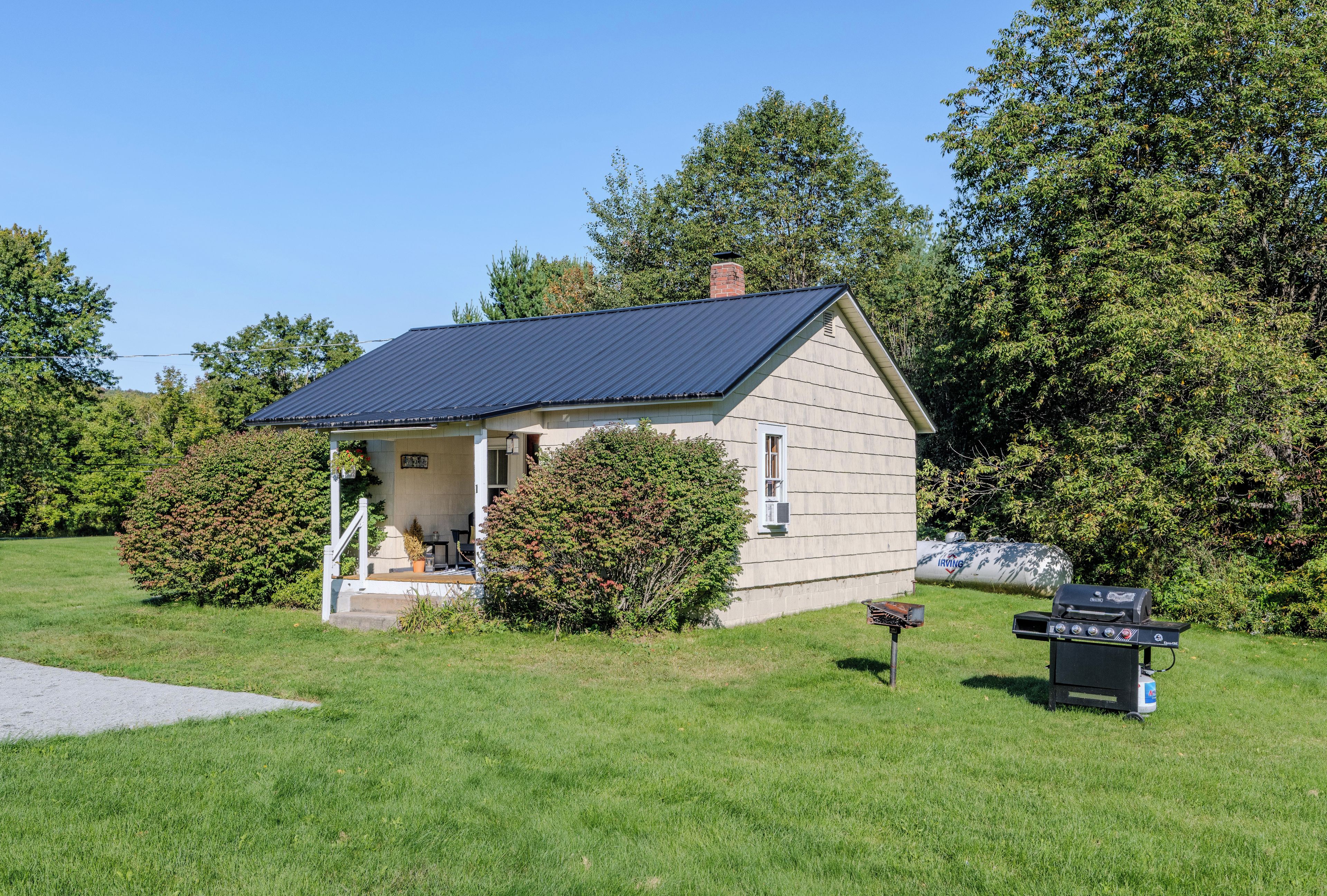Een klein, gelijkvloers huis met een schuin dak, een veranda en een barbecue in de tuin.