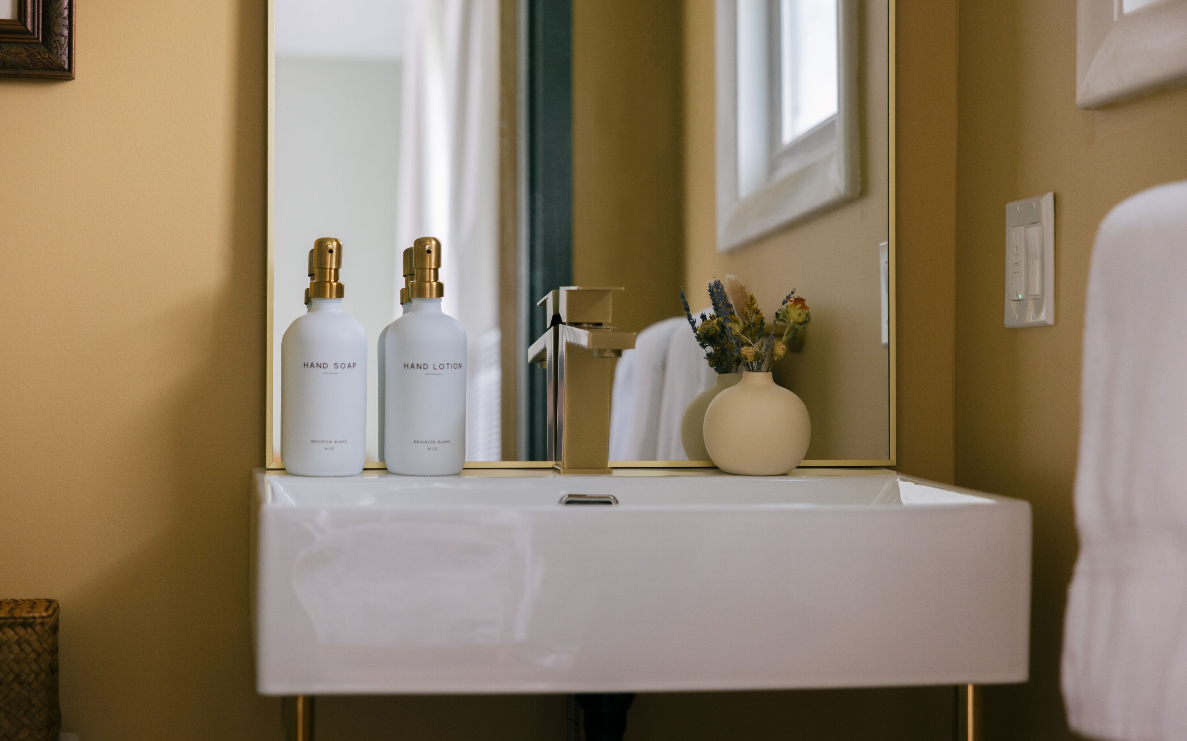 Une salle de bain avec un lavabo blanc, deux flacons de savon pour les mains et de lotion, un robinet et un vase avec des fleurs.