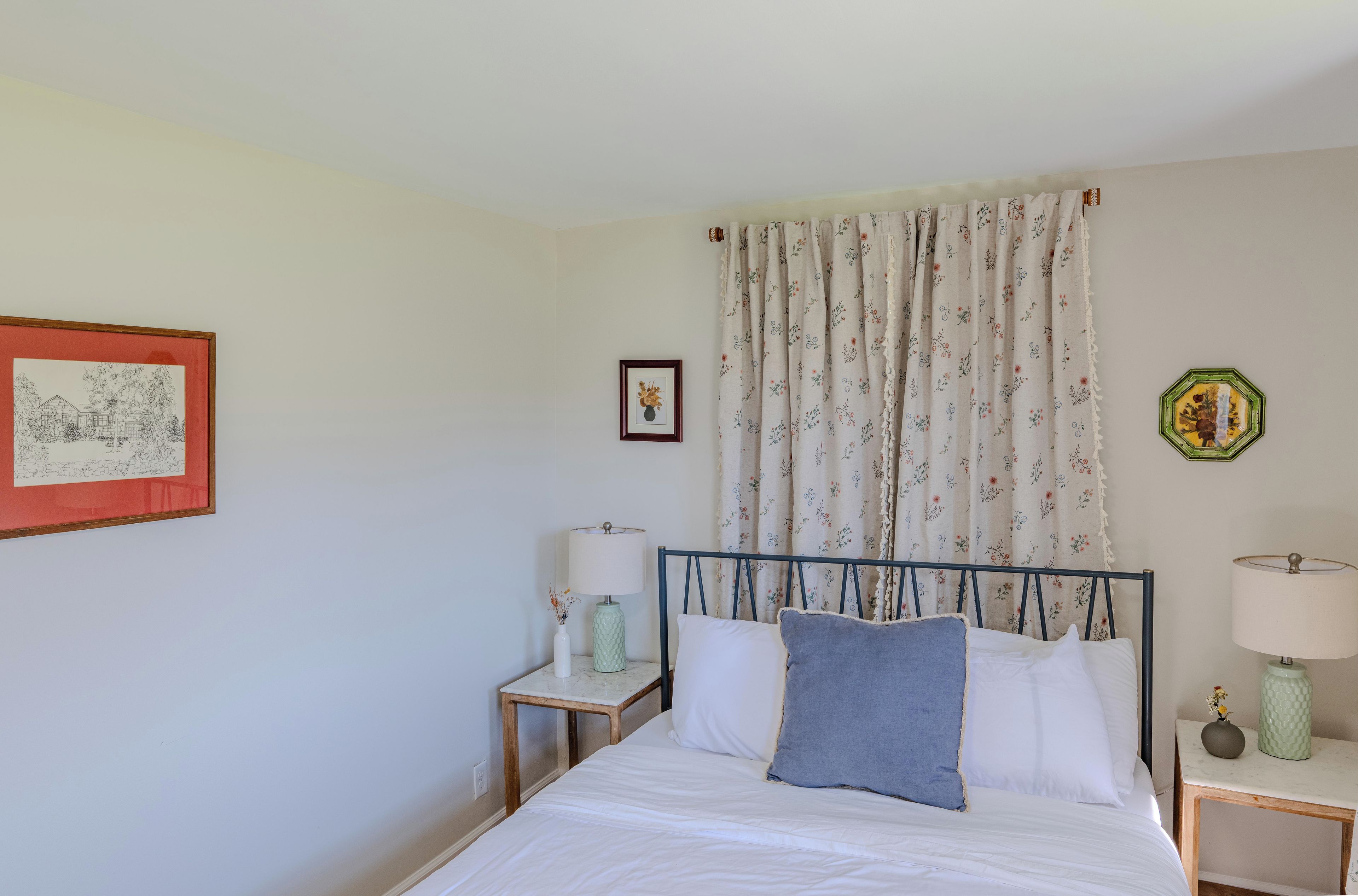 Un dormitorio con cama, mesita de noche, lámpara, un cuadro enmarcado y cortina con estampado floral.