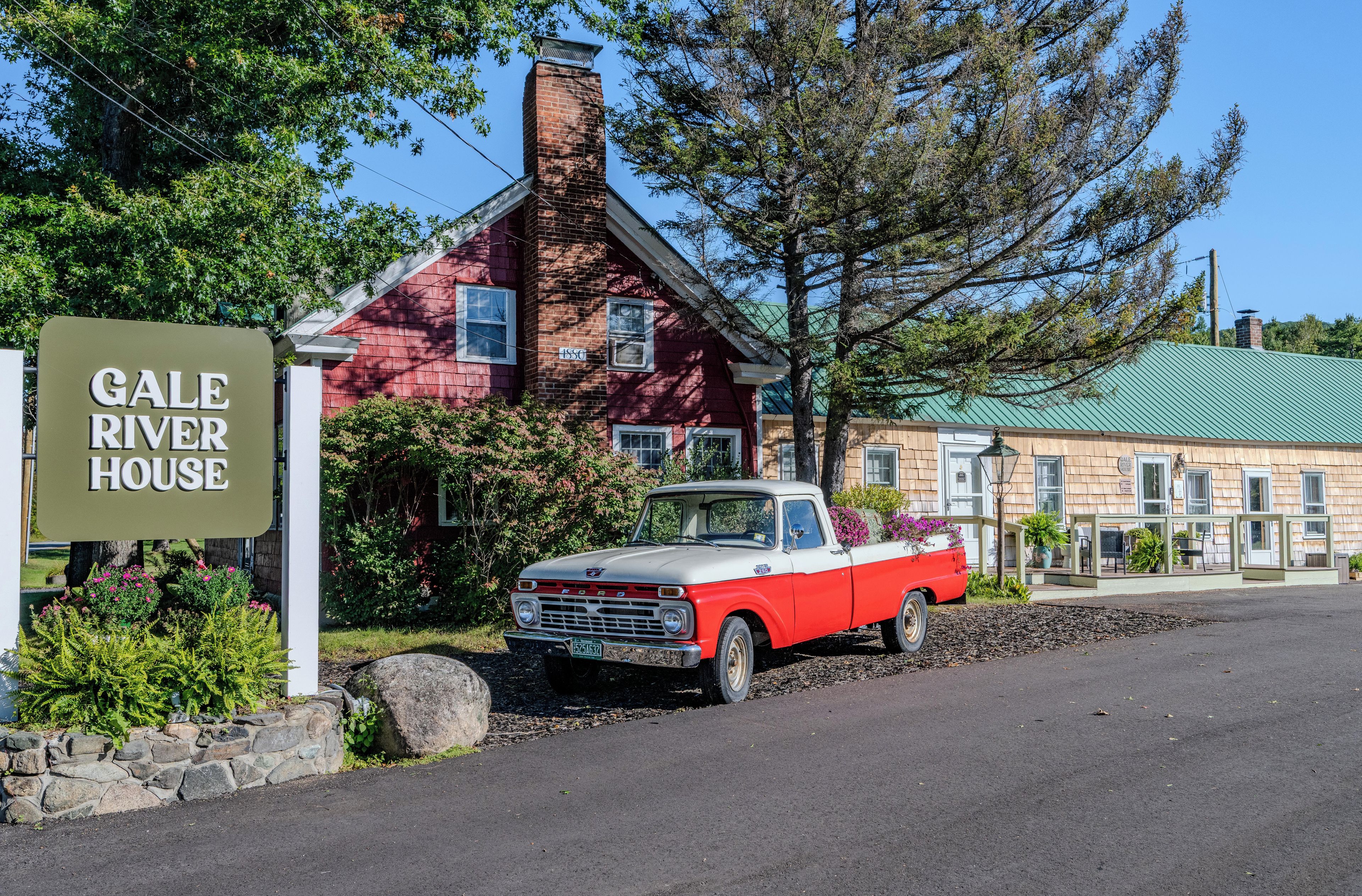 Un camion Ford ancien, rouge et blanc, est garé devant une maison en briques rouges surmontée d’un toit vert, et une plaque indique : « Gale River House ».