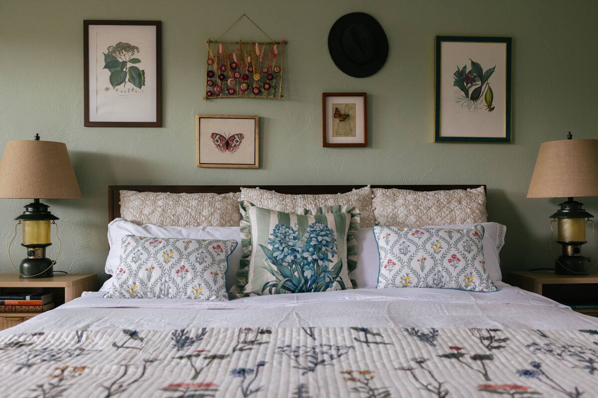 A neatly made bed with floral-patterned pillows, a headboard with a decorative panel, and framed botanical illustrations on the wall.
