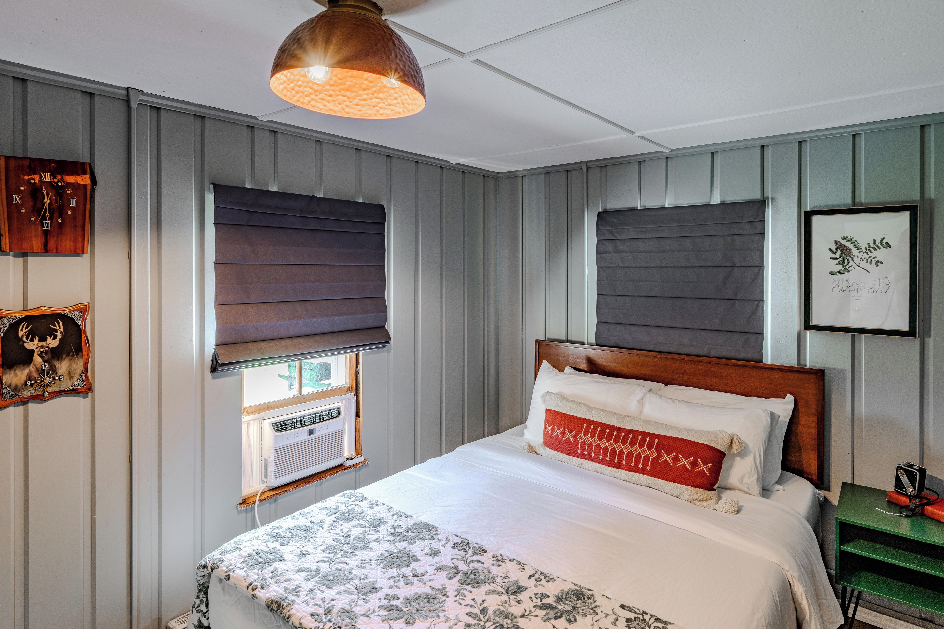 Un dormitorio con cama, una ventana con persianas, un reloj de pared e impresión enmarcadas de motivos botánicos.