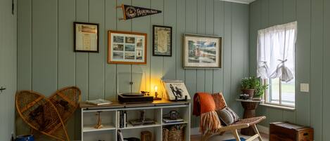 Una habitación con una estantería, cuadros enmarcados, una lámpara, una silla y una ventana con cortinas.
