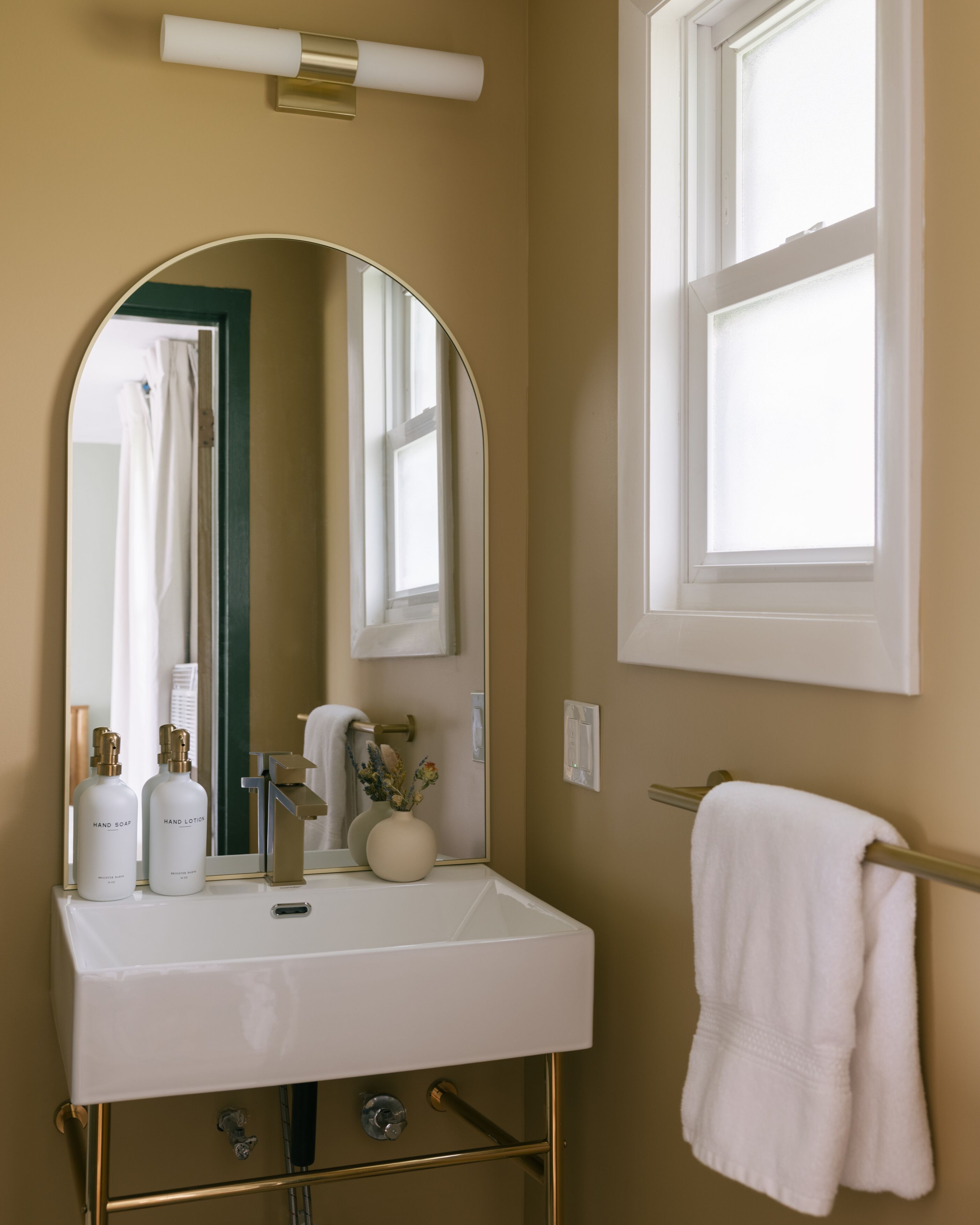 A bathroom with a white sink, gold fixtures, a mirror, a window, and a towel rack.
