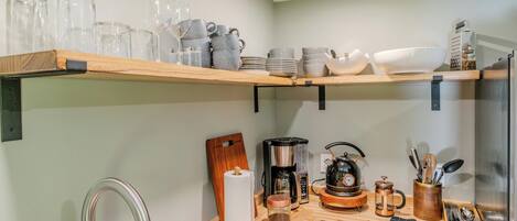 A kitchen with wooden shelves holding glassware and dishes, a coffee maker, a kettle, and a knife block.