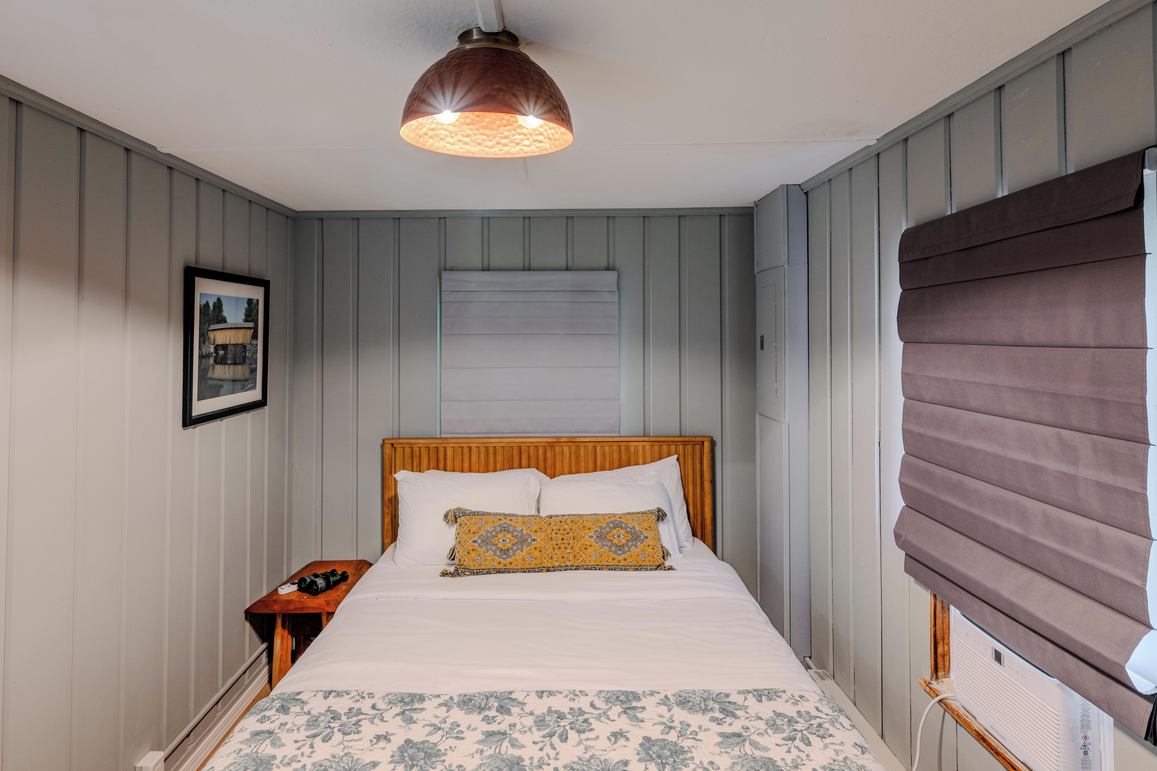 Un dormitorio con cabecera de madera, cama con ropa de cama blanca y un cojín decorativo estampado, mesita de noche con lámpara y ventana con persianas.