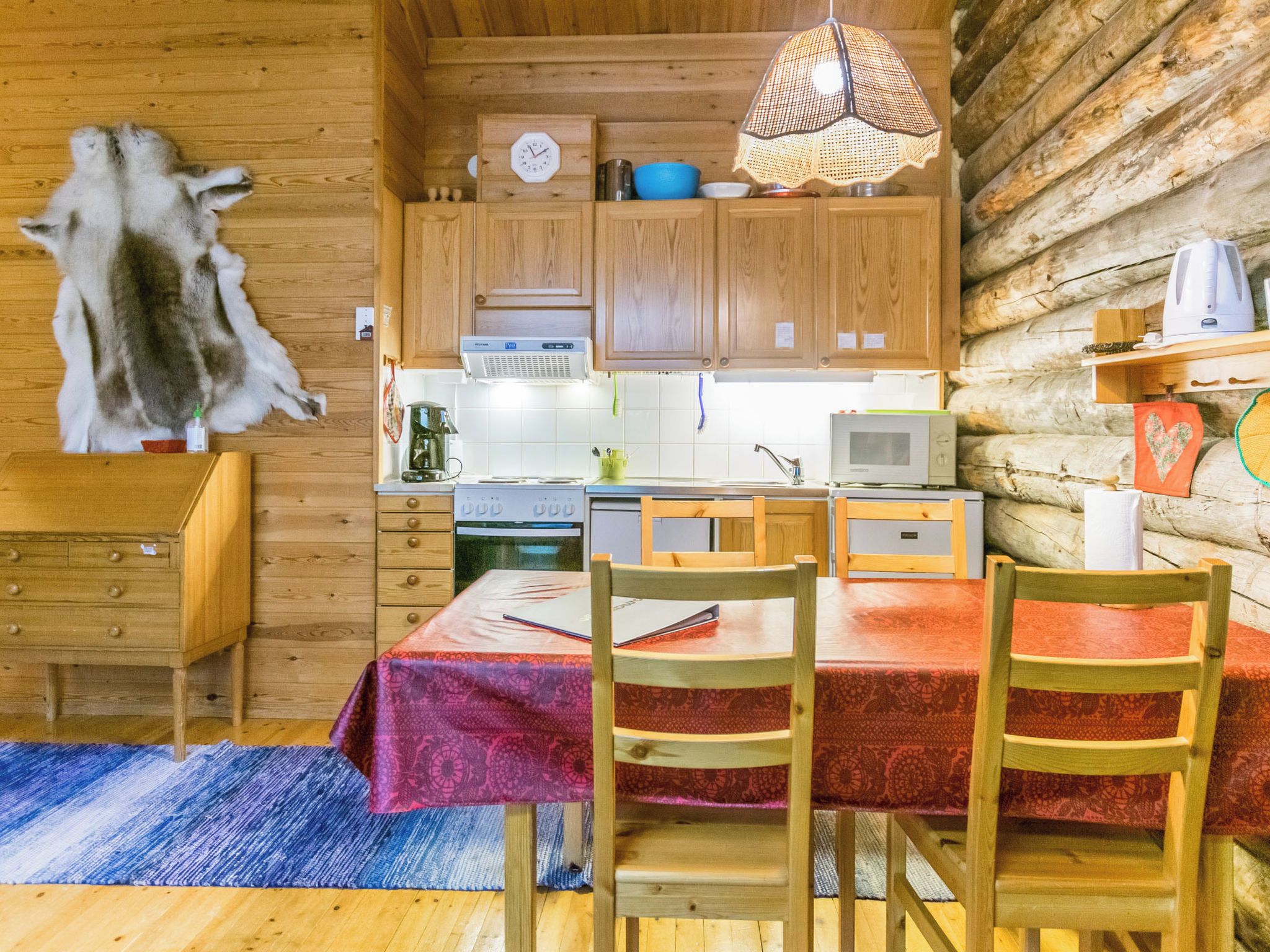 A cabin interior with a wooden dining table, chairs, a kitchen area with appliances, and a hanging light fixture.