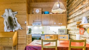 Interior rústico de una cabaña con paredes de madera, una mesa de comedor con sillas, una zona de cocina con electrodomésticos y una lámpara colgante.