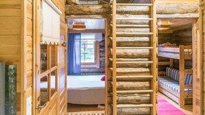 Interior de una cabaña de troncos con cama en un altillo, escalera y literas.