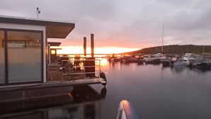 En småbåtshamn med förtöjda båtar, en modern byggnad med stora fönster och en solnedgång i bakgrunden.