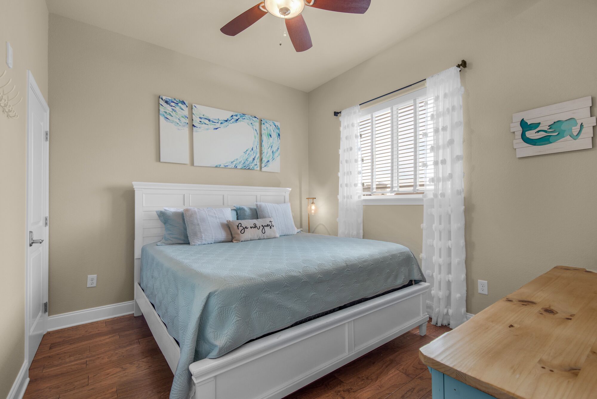 A bedroom with a bed, a ceiling fan, a window with white curtains, and artwork on the walls.