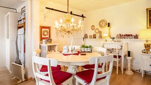 Un comedor con una mesa redonda, sillas blancas con cojines rojos, una araña y una chimenea blanca decorada con objetos.