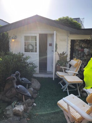 Una casita con la puerta abierta, una ventana y un letrero en la pared. Hay dos sillas afuera y una formación rocosa con dos pájaros.
