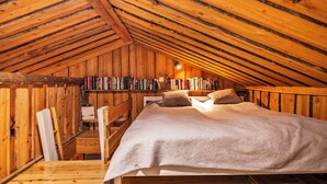 Ein gemütliches Schlafzimmer im Dachgeschoss mit Holzbalkendecke, einem Bett mit Kissen, einem Schreibtisch mit Stuhl und einem Bücherregal voller Bücher.