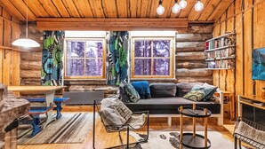 Ein gemütliches Blockhaus-Interieur mit Holzbalkendecke, einer Couch mit Kissen, einem kleinen runden Tisch, einem Bücherregal und einem Fenster mit Vorhängen.