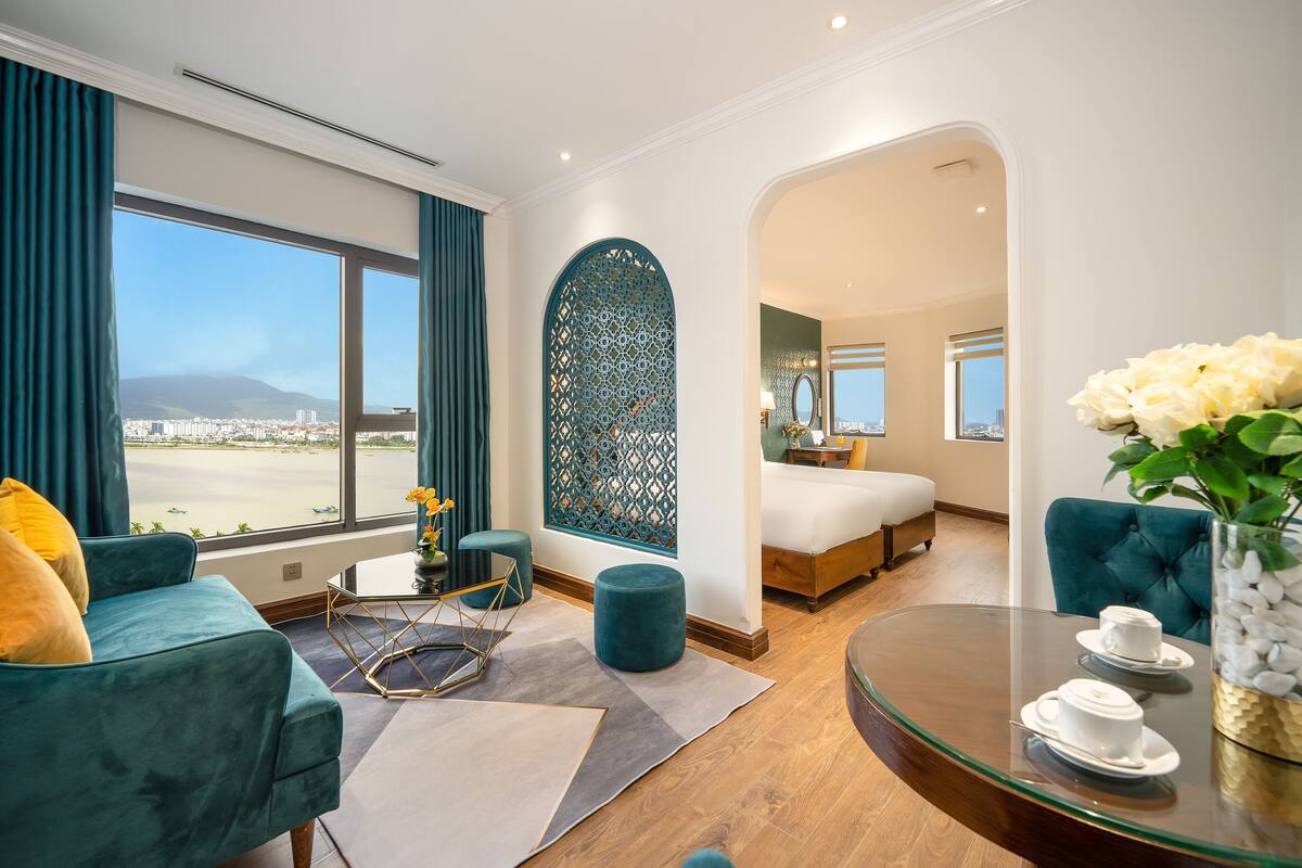 Une chambre d’hôtel moderne dotée d’une grande fenêtre, d’un canapé recouvert d’un tissu turquoise, d’une table basse en verre et d’un lit offrant une vue sur la ville.