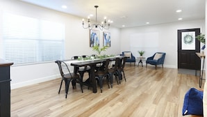 A dining area with a wooden table, chairs, and a chandelier. There are two windows with blinds, a door with a wreath, and a small plant on a side table. - Champions Getaway (San Antonio)