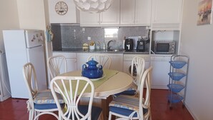 A compact kitchen with white cabinets, a round dining table with chairs, a blue pot, and a blue basket on a stand. - Varandas da Rocha 7 (Portimão)